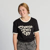A person with light brown hair in a ponytail smiles while wearing the New Duds Cheese the Day Ringer T-shirt in black and cream. They are also wearing gray jeans, and the background is plain light gray.