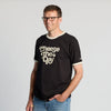 A person wearing glasses and a New Duds Cheese the Day Ringer T-shirt in Black & Cream stands smiling with one hand in their jeans pocket against a plain background.
