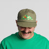 A man with a mustache is wearing a green shirt and the New Duds Fresh From Vermont 90s Roper Hat—an olive cap featuring a mountain and tent design—as he looks down and smiles against a plain light background.