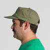A man with a mustache, wearing a green t-shirt and the Fresh From Vermont 90s Roper Hat by New Duds—an olive green cap featuring “CABOT” embroidered on the side—is shown in profile against a plain light background.