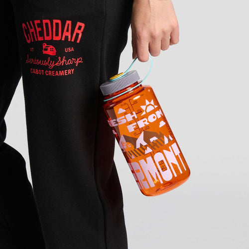 A person wearing black pants with “CHEDDAR” text holds a Downeast Bicycle Specialists Fresh From Vermont Nalgene water bottle by its strap.