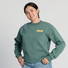 A person wearing the New Duds “Life’s Better With Butter” crewneck, featuring a green sweatshirt with a yellow BUTTER patch, and light blue jeans smiles with hands on hips against a plain light background.