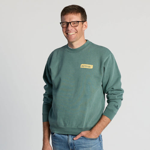A smiling man in glasses stands against a plain light background with one hand in his pocket, wearing blue jeans and the “Life’s Better With Butter Crewneck” by New Duds.