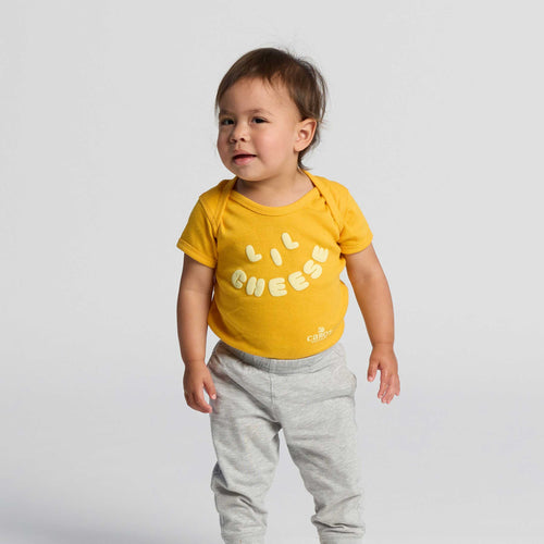 A toddler wearing the New Duds Lil Cheese Onesie in yellow and light gray pants stands against a plain white background, looking slightly to the side.