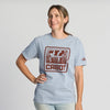 A woman smiles and poses in the Vintage Cabot Cow Graphic T-shirt by New Duds, featuring a red CABOT logo with cows on light blue fabric. She stands against a plain white background, one arm on her hip and the other relaxed.