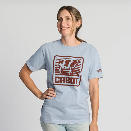 A woman smiles and poses in the Vintage Cabot Cow Graphic T-shirt by New Duds, featuring a red CABOT logo with cows on light blue fabric. She stands against a plain white background, one arm on her hip and the other relaxed.