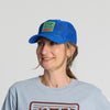 A woman is smiling and looking slightly to the side, wearing the New Duds Vintage Cabot Cow Graphic Mesh Trucker Hat—a blue mesh cap with a green and red patch—along with a light blue graphic T-shirt and gold hoop earrings.