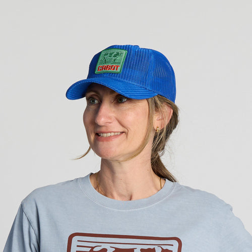 A woman is smiling and looking slightly to the side, wearing the New Duds Vintage Cabot Cow Graphic Mesh Trucker Hat—a blue mesh cap with a green and red patch—along with a light blue graphic T-shirt and gold hoop earrings.