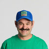 A man with a mustache, dressed in a green t-shirt and smiling at the camera against a plain light background, wears the New Duds Vintage Cabot Cow Graphic Mesh Trucker Hat—a blue trucker hat with a mesh crown and accented with a green and red patch.