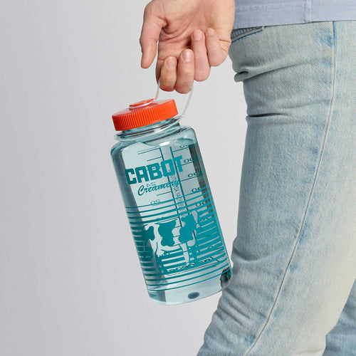 A person in light blue jeans holds the Cabot Cow Nalgene Water Bottle by Downeast Bicycle Specialists. The clear bottle, filled with water, features a red cap, blue “CABOT” branding, cow silhouettes, and measurement markings.