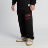 A person wearing New Duds Seriously Sharp Sweatpants in black, featuring CHEDDAR and red text printed on the upper thigh, paired with a black sweatshirt and beige sneakers with a green N logo, stands against a plain white background.