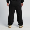 A person stands facing away, wearing New Duds Seriously Sharp Sweatpants in black, along with a black sweatshirt and beige and white sneakers. The background is plain and light-colored.