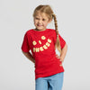 A young child with long, light brown braided hair smiles while standing with hands behind their back. The child is wearing the New Duds Big Cheese Toddler T-shirt in red, made from soft ringspun cotton and featuring yellow “BIG CHEESE” lettering, paired with light blue pants against a plain background.