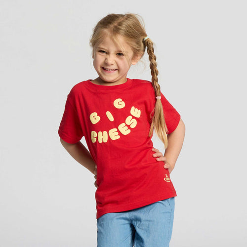 A young child with long, light brown braided hair smiles while standing with hands behind their back. The child is wearing the New Duds Big Cheese Toddler T-shirt in red, made from soft ringspun cotton and featuring yellow “BIG CHEESE” lettering, paired with light blue pants against a plain background.