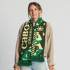 A woman with light brown hair, dressed in a tan jacket and jeans, smiles at the camera. She has the Fresh From Vermont Scarf by Ruffneck Wear, INC. draped around her neck, its green knit fabric and farm-themed designs adding a touch of local charm to her outfit.