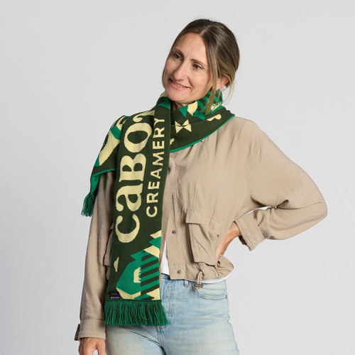 A woman in a beige shirt and light jeans smiles while posing with the Fresh From Vermont Scarf by Ruffneck Wear, INC.—a green knit accessory proudly displaying “CABOT CREAMERY”—against a plain white background, showcasing her Vermont pride.
