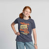 A smiling girl with red hair in pigtails wears the New Duds Seriously Sharp Riddle Youth T-shirt, featuring the phrase “Which cheese is the smartest?” and an illustration of Cabot Seriously Sharp Cheddar, as she stands against a plain light background.