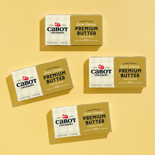 Four sticks of Cabot Creamery Baker’s Butter (Unsalted 4 Pack) are arranged diagonally on a bright yellow background—two labeled unsalted in gold, two extra creamy in dark gold, perfect for baking or pastries.