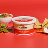 A Cabot Creamery Sour Cream container rests on a red surface, perfect for a family dinner. Triangular tortilla chips, one dipped in the creamy staple, accompany a salsa bowl garnished with cilantro.