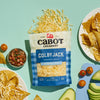 A bag of Cabot Creamery Colby Jack Shredded Cheese sits on a light blue background, surrounded by tortilla chips with cheese, sliced avocado, cherry tomatoes, and a bowl of salsa.