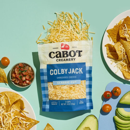 A bag of Cabot Creamery Colby Jack Shredded Cheese sits on a light blue background, surrounded by tortilla chips with cheese, sliced avocado, cherry tomatoes, and a bowl of salsa.