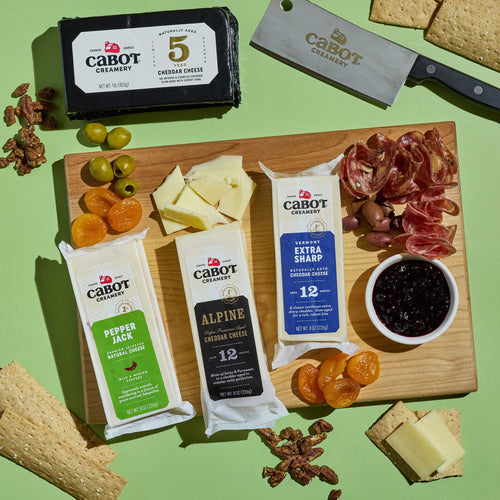 A Cabot X That Cheese Plate: Ultimate Charcuterie Gift Box on a wooden board featuring Cabot Creamery cheeses, crackers, salami, olives, dried apricots, a bowl of jam, a knife, and an additional block of aged cheddar.