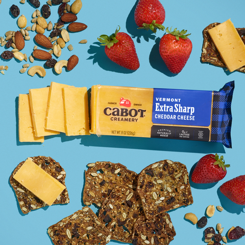 A block of Cabot Creamery Extra Sharp Yellow Cheddar Cheese sits with slices, surrounded by strawberries, multigrain crackers, almonds, and dried fruit on a bright blue background.