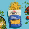 An open bag of Cabot Creamery Extra Sharp Yellow Cheddar Cheese spills shredded cheese onto a blue surface, surrounded by arugula, sliced radish, cherry tomatoes, and a fresh salad.