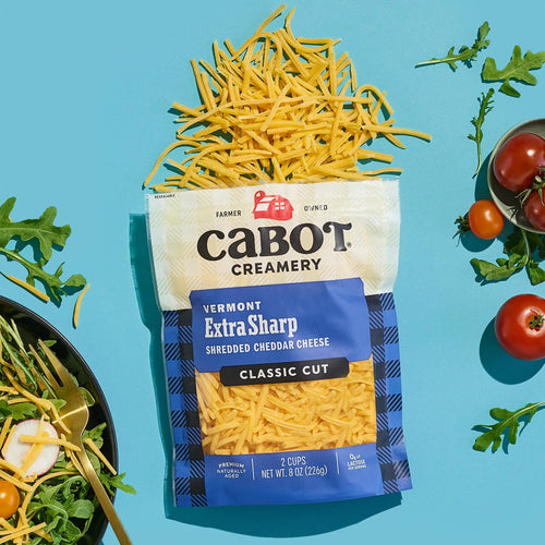 An open bag of Cabot Creamery Extra Sharp Yellow Cheddar Cheese spills shredded cheese onto a blue surface, surrounded by arugula, sliced radish, cherry tomatoes, and a fresh salad.