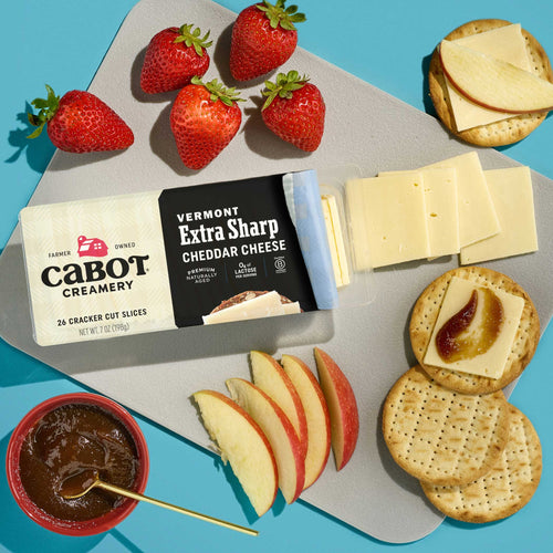 A tray featuring Cabot Creamery Extra Sharp Cheddar Cheese slices, crackers with cheddar and jam, sliced apples, strawberries, and a bowl of jam is arranged on a light gray surface in bright lighting.