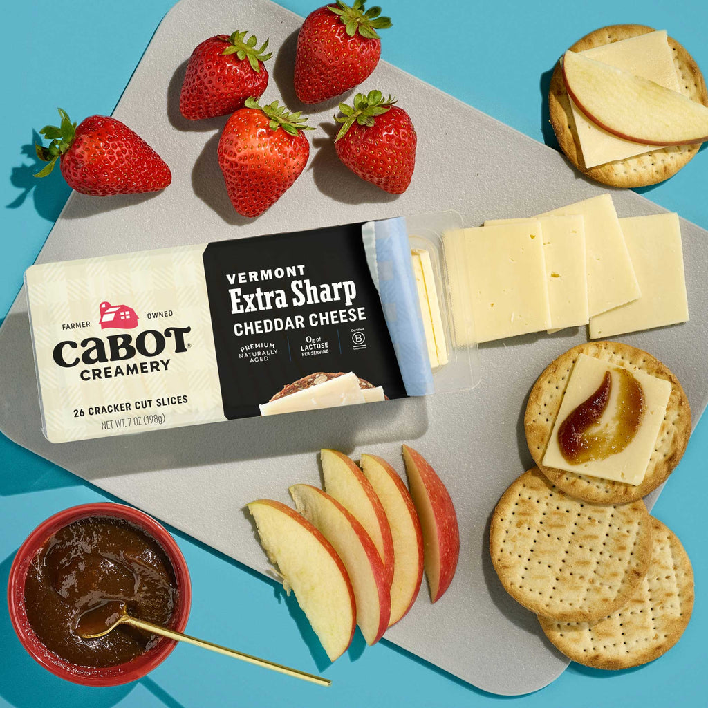 A tray featuring Cabot Creamery Extra Sharp Cheddar Cheese slices, crackers with cheddar and jam, sliced apples, strawberries, and a bowl of jam is arranged on a light gray surface in bright lighting.