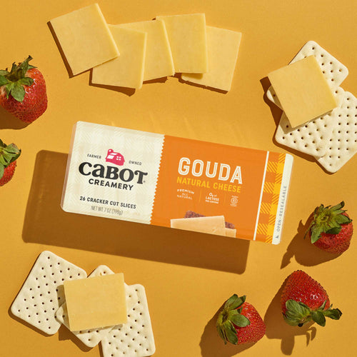 Cabot Creamery Gouda Cheese slices are paired with square crackers and fresh strawberries on a yellow surface, with some cheese slices set atop the crackers.