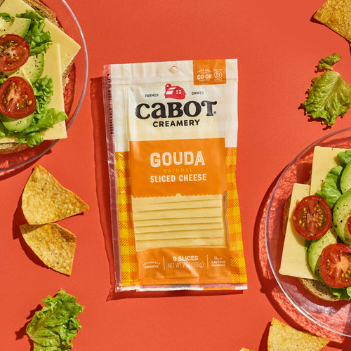 A package of Cabot Creamery Gouda Cheese sits on an orange surface, surrounded by tortilla chips and plates with sandwiches topped with avocado, tomato, lettuce, and melted cheese.