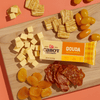 A wooden board with cubes of Cabot Creamery Gouda Cheese, its packaging, stacked crackers, yellow grape tomatoes, dried apricots, and sliced cured meat forms a colorful, protein-rich snack on an orange background.