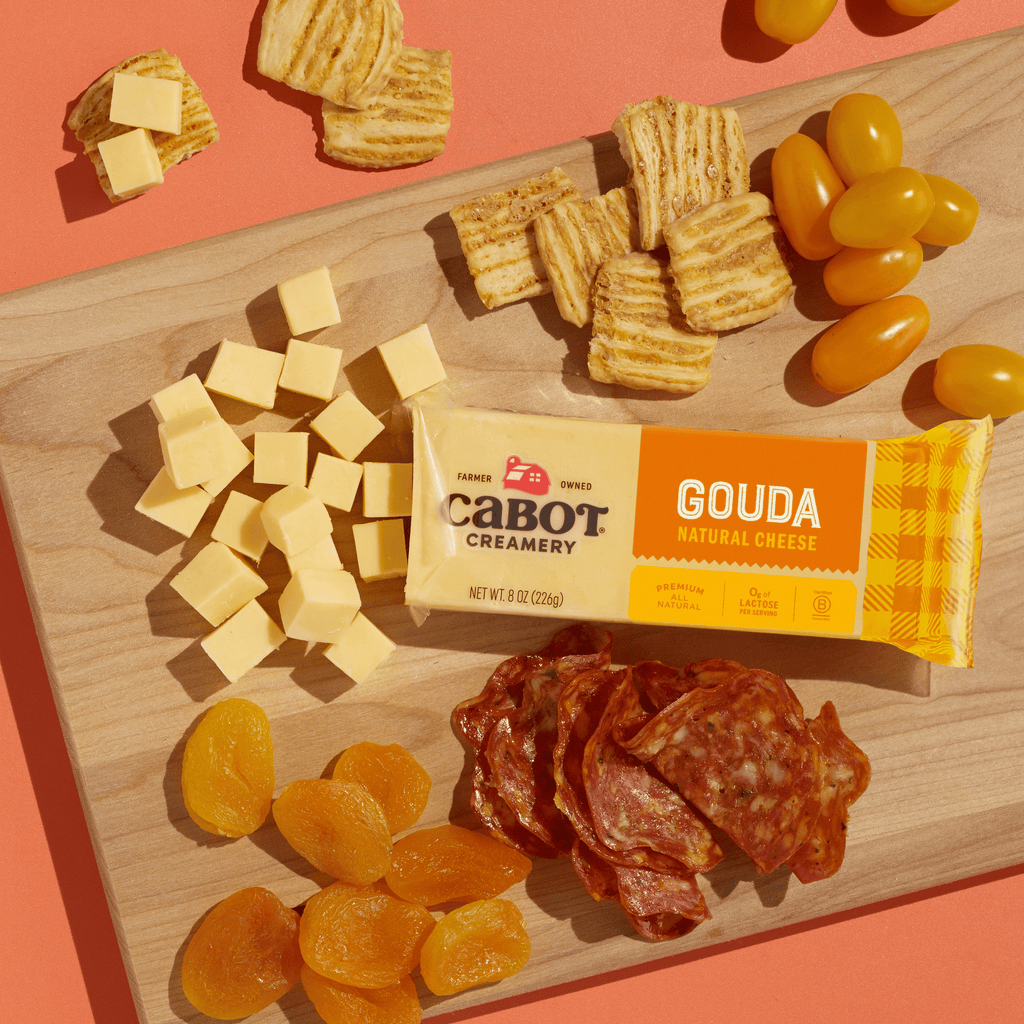 A wooden board with cubes of Cabot Creamery Gouda Cheese, its packaging, stacked crackers, yellow grape tomatoes, dried apricots, and sliced cured meat forms a colorful, protein-rich snack on an orange background.