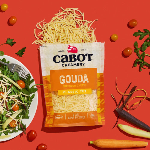 A package of Cabot Creamery Gouda Cheese sits on a red background with shredded cheese spilling out, surrounded by salad greens, grape tomatoes, a fresh plate of salad, and colorful carrots.