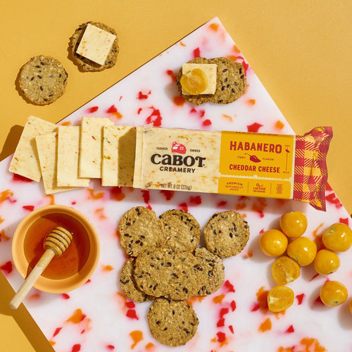 A block and slices of Cabot Creamery Habanero Cheddar Cheese are arranged on a marble board with crackers, honey, and yellow cherry tomatoes, all set against a yellow background.