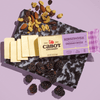 A partially sliced block of Cabot Creamery Horseradish Cheddar Cheese rests on a black and white marbled board with mixed nuts and fresh blackberries, set against a light purple background.
