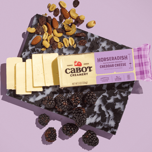 A partially sliced block of Cabot Creamery Horseradish Cheddar Cheese rests on a black and white marbled board with mixed nuts and fresh blackberries, set against a light purple background.