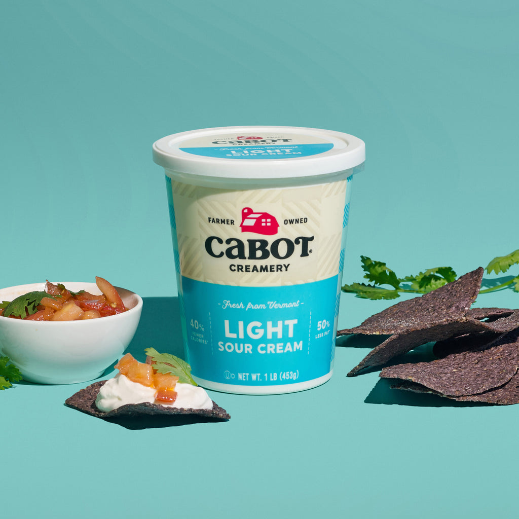 A container of Cabot Creamery Lite Sour Cream, with 50% less fat, rests on a turquoise background surrounded by blue corn tortilla chips and a small bowl of salsa, with scattered cilantro leaves.