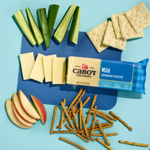 Cabot Creamery Mild Cheddar Cheese, sliced and lactose free, is served with cucumber sticks, gluten-free crackers, pretzel sticks, and apple slices on a blue background.