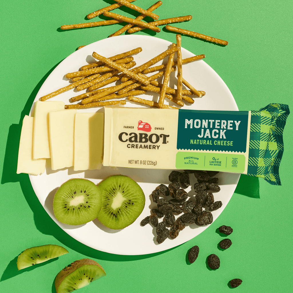 On a green background, a white plate holds Cabot Creamery Monterey Jack Cheese slices, pretzel sticks, kiwi slices, raisins, and a partially unwrapped block of Cabot Creamery Monterey Jack Cheese.