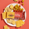 A plate with Cabot Creamery New York Extra Sharp Yellow Cheddar Cheese, square crackers, and red and yellow grape tomatoes sits on an orange surface. Some cheddar, crackers, and tomatoes are scattered around the plate.