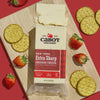 A block of Cabot Creamery New York Extra Sharp Cheddar Cheese rests on a wooden board, surrounded by round crackers and whole strawberries, against a red background.