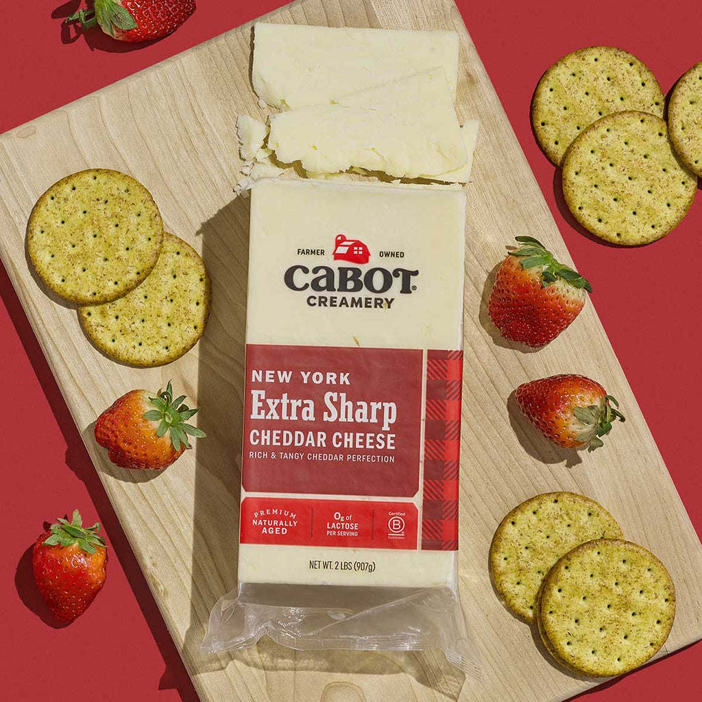 A block of Cabot Creamery New York Extra Sharp Cheddar Cheese rests on a wooden board, surrounded by round crackers and whole strawberries, against a red background.