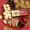 On a vibrant red background, a round wooden board displays Cabot Creamery’s New York Extra Sharp Cheddar Cheese cubes, whole grain crackers, fresh blackberries, red pear slices, a whole red pear, and the cheese package.