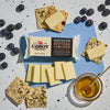 A block of Cabot Creamerys lactose-free New York Vintage Cheddar Cheese is elegantly arranged with cheese slices on herb crackers, fresh blueberries, and a honey jar with dipper on a speckled surface.