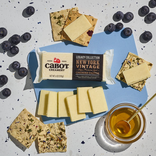 A block of Cabot Creamerys lactose-free New York Vintage Cheddar Cheese is elegantly arranged with cheese slices on herb crackers, fresh blueberries, and a honey jar with dipper on a speckled surface.