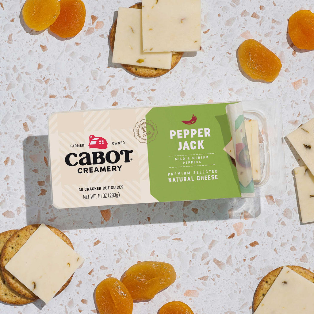 Packaged Cabot Creamery Pepper Jack cheese on a terrazzo countertop with crackers and dried apricots.
