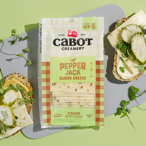 On a green background, a package of Cabot Creamerys Pepper Jack Cheese showcases two open sandwiches with slices, pickles, and greens. This creamy treat is spiced up with jalapenos and is clearly labeled gluten-free.